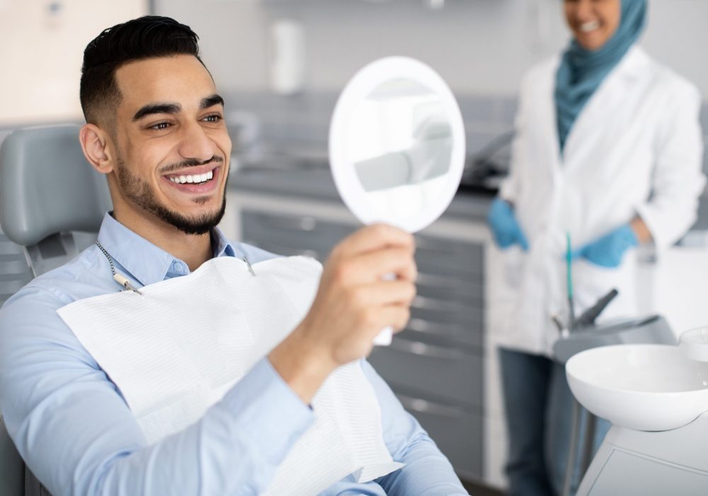 Happy Handsome Arab Man Looking To Mirror After Teeth Treatment In Modern Clinic, Middle Eastern Male Patient Sitting In Chair In Stomatological Cabinet And Enjoying His New Smile, Closeup